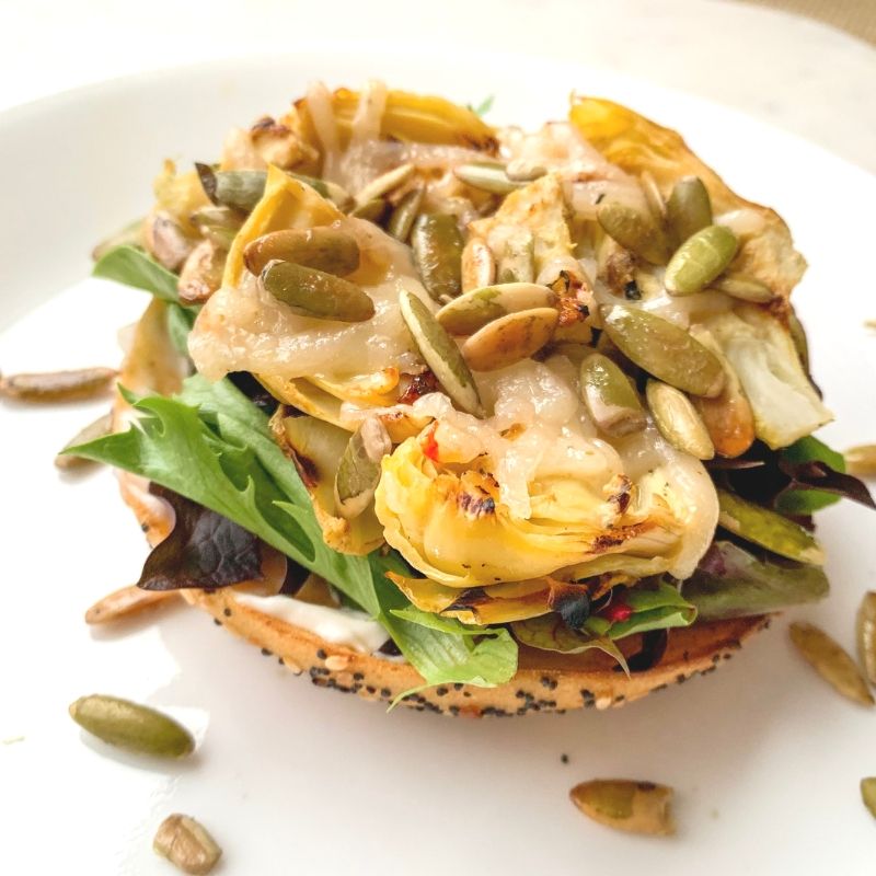 side view bagel with mixed greens, grilled artichoke hearts, broiled vegan cheese, raw pumpkin seeds